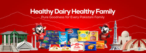 A promotional banner with a bright red background featuring the text 'Healthy Dairy Healthy Family' and 'Pure Goodness for Every Pakistani Family.' Below the text is a grouped arrangement of packaged food products, including milk, butter, biscuits, and other dairy items, flanked by cartoon cow characters and Pakistani landmarks such as the Taj Mahal and Red Fort.